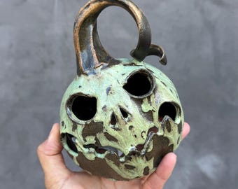 May include: A ceramic pumpkin-shaped lantern featuring a dark brown base with a distressed light green glaze overlay. The piece has a curved stem handle and carved openings for eyes, a nose, and a mouth, creating a hollow interior structure.