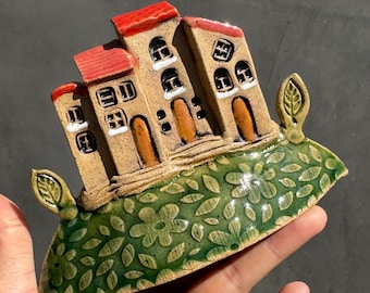 May include: A ceramic sculpture of a small village with red roofs and green foliage. The houses are detailed with windows and doors, and the base is decorated with a floral pattern.