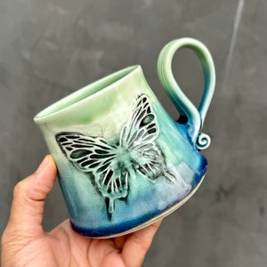 Può includere: Una tazza in ceramica con una glassa ombré blu, verde e bianca. La tazza presenta un design a farfalla in rilievo in nero.