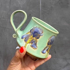 May include: A handmade ceramic mug with a pale green glaze. The mug features blue and yellow iris flower designs. The handle is a loop shape. The mug is held by a person with red nail polish.
