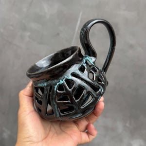 Puede incluir: Una taza de cerámica negra con un diseño calado único. La taza presenta un asa curvada y un acabado brillante con detalles turquesa. El patrón calado crea un efecto decorativo, ideal para bebidas.