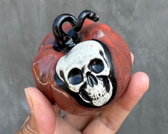 May include: A small, round, reddish-brown ceramic pumpkin ornament featuring a detailed white skull centered on the front. The pumpkin has a glossy finish and a curved, black, high-gloss stem extending from the top center of the object.