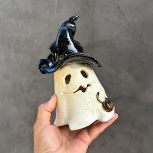 May include: A ceramic ghost figurine with a speckled off-white body and a glossy black witch hat. The ghost features cut-out eyes and a mouth, with a small brown pumpkin held in one hand. The surface has a smooth, glazed finish throughout.