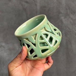 May include: A light green ceramic bowl with a lattice design. The bowl has a rounded shape with cut-out geometric patterns. The bowl is held in a hand against a gray background. The bowl is glazed with a glossy finish.