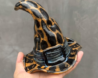 May include: A ceramic witch hat sculpture featuring a glossy finish with a black and orange mottled pattern. The hat has a pointed, curved top and a wide brim, accented by a dark blue band around the base that includes a rectangular buckle detail.