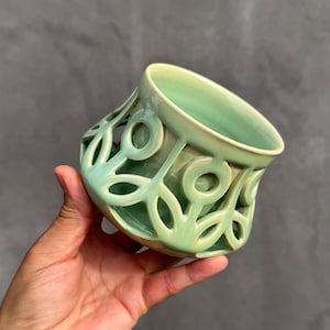 Puede incluir: Un cuenco de cerámica verde claro con un diseño calado. El cuenco se sostiene en una mano y tiene una forma acampanada y un acabado liso y brillante. El patrón recortado crea un motivo floral decorativo.