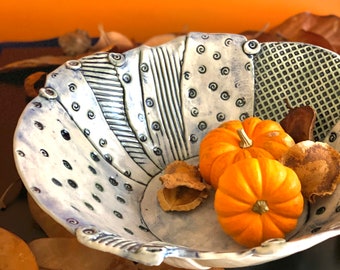 May include: A handcrafted ceramic bowl with a white and blue design, featuring patterns of lines, dots, and squares. The bowl holds three small orange pumpkins and dried leaves, creating a fall harvest theme.