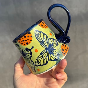 May include: A yellow ceramic mug with a blue handle and a butterfly design. The mug is decorated with orange and black polka dots.