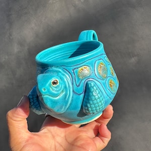 May include: A turquoise ceramic mug in the shape of a turtle. The mug has a detailed turtle face, flipper-like handles, and a shell design with brown and yellow accents. The mug is held in a hand.