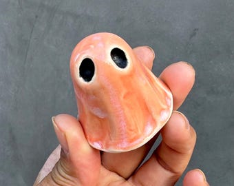 May include: A small, glossy, peach-colored ceramic figurine shaped like a ghost. The object features a smooth, rounded top and a flared base with two deep, dark, oval-shaped indentations serving as eyes, creating a simple, hollow-eyed expression.