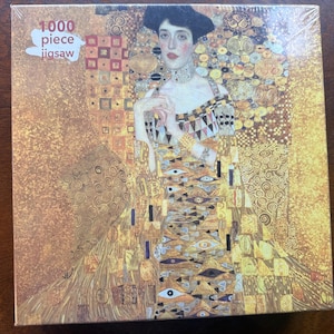 Puede incluir: Un rompecabezas de 1000 piezas que representa una pintura de una mujer con un vestido dorado con un patrón geométrico. El fondo es un mosaico dorado y marrón.