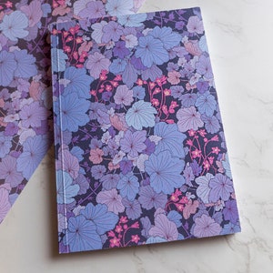 May include: Two notebooks with a floral pattern in shades of purple, pink, and blue. The notebooks are lying on a white marble surface.