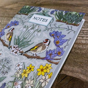 May include: A gray notebook with a floral and bird design. The cover features a variety of flowers, including daffodils, snowdrops, and crocuses, as well as two goldfinches. The title "NOTES" is printed in a white rectangle on the cover.