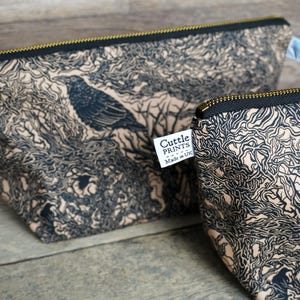 May include: Two beige and black zippered pouches with a crow and tree branch design. The pouches have a black zipper and a gold-colored zipper pull. The label on the pouch reads "Cuttle Prints Made in UK".