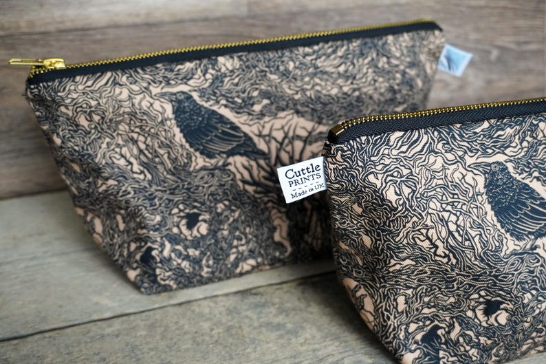 May include: Two beige and black zippered pouches with a crow and tree branch design. The pouches have a black zipper and a gold-colored zipper pull. The label on the pouch reads "Cuttle Prints Made in UK".