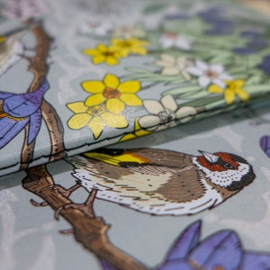 May include: A close-up of a fabric with a bird and floral pattern. The bird is brown and white with a yellow beak. The flowers are yellow, white, and purple. The fabric is a light gray color.