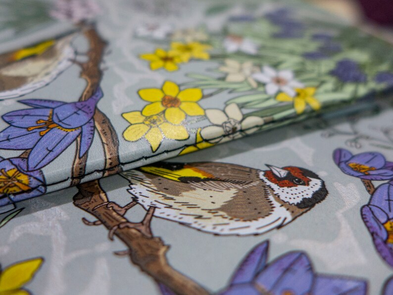 May include: A close-up of a fabric with a bird and floral pattern. The bird is brown and white with a yellow beak. The flowers are yellow, white, and purple. The fabric is a light gray color.