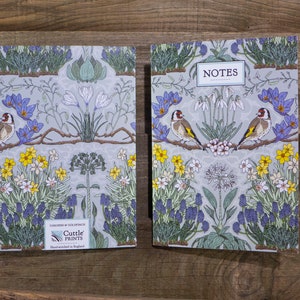 May include: A pair of gray and white floral patterned notebooks with a bird design. The front cover of the notebook on the left has the text "Cuttle Prints" and "Gardens of Goldfinch" printed on it. The front cover of the notebook on the right has the text "NOTES" printed on it.