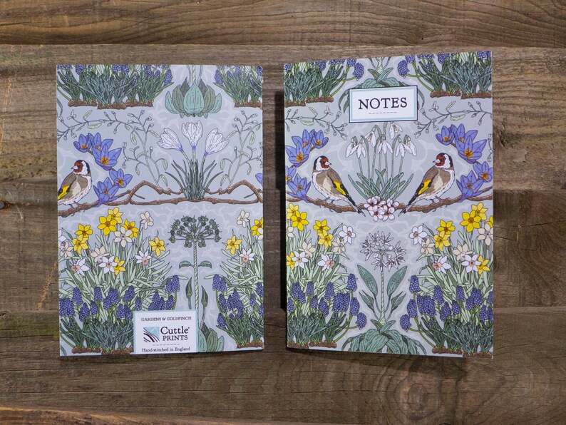 May include: A pair of gray and white floral patterned notebooks with a bird design. The front cover of the notebook on the left has the text "Cuttle Prints" and "Gardens of Goldfinch" printed on it. The front cover of the notebook on the right has the text "NOTES" printed on it.