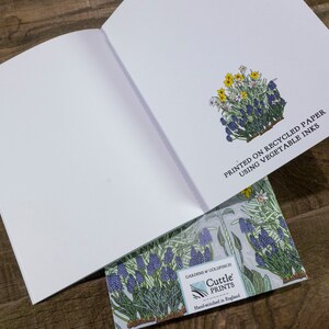 May include: An open blank notebook with a floral design on the back cover. The back cover features a cluster of blue and yellow flowers with green leaves. The text "PRINTED ON RECYCLED PAPER USING VEGETABLE INKS" is printed on the back cover. The text "GARDENS & GOLDFINCH Cuttle PRINTS Hand-wished in England" is printed on the bottom of the back cover.