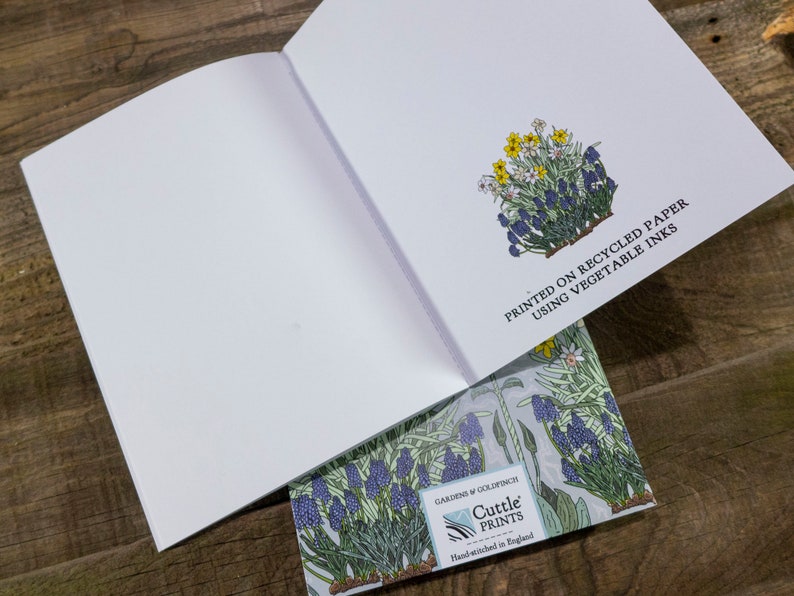 May include: An open blank notebook with a floral design on the back cover. The back cover features a cluster of blue and yellow flowers with green leaves. The text "PRINTED ON RECYCLED PAPER USING VEGETABLE INKS" is printed on the back cover. The text "GARDENS & GOLDFINCH Cuttle PRINTS Hand-wished in England" is printed on the bottom of the back cover.
