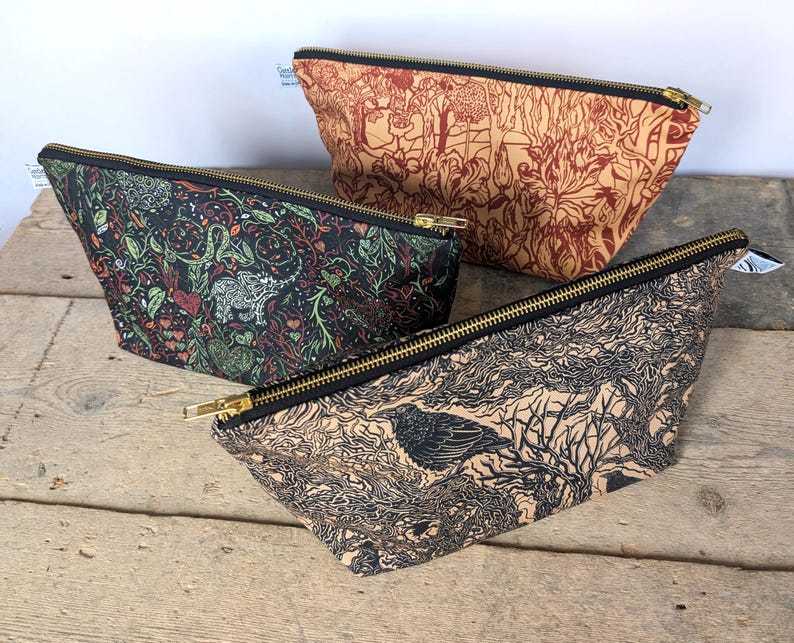 May include: Three cosmetic bags with black zippers and patterned fabric. One bag has a black background with a floral design, another has a tan background with a botanical print, and the third has a tan background with a bird and tree design.