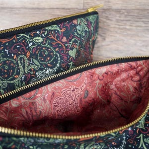 May include: Two zippered pouches with a black exterior featuring a green, orange, and white botanical pattern. The interior lining is a coral color with a white elephant and floral design. Gold-colored zippers.