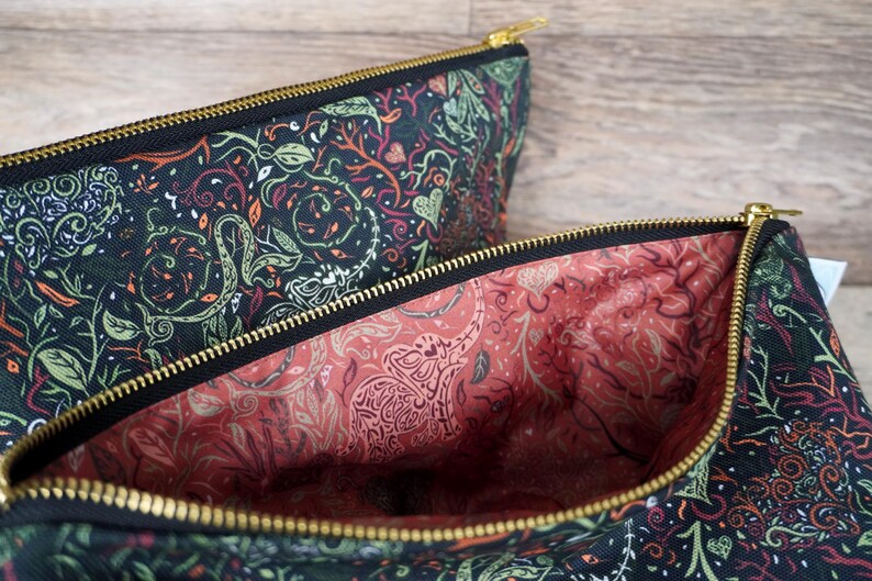May include: Two zippered pouches with a black exterior featuring a green, orange, and white botanical pattern. The interior lining is a coral color with a white elephant and floral design. Gold-colored zippers.