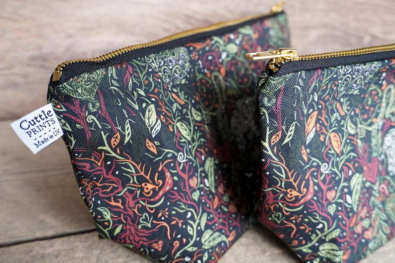 May include: Two zippered pouches with a dark background and a colorful botanical pattern. The design includes green, orange, and red plant elements. The pouches have gold-colored zippers and a white tag that reads "Cuttle Prints - Made in UK."