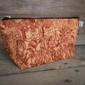 May include: A rectangular cosmetic bag with a floral pattern in shades of rust and tan. The bag has a black zipper and a small white tag. The bag is made of fabric and sits on a wooden surface.