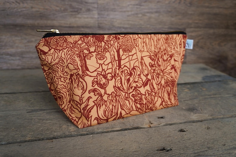 May include: A rectangular cosmetic bag with a floral pattern in shades of rust and tan. The bag has a black zipper and a small white tag. The bag is made of fabric and sits on a wooden surface.
