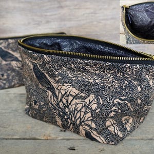 May include: Two fabric cosmetic bags with a black and tan tree and bird design. The bags have a black zipper with a gold-colored zipper pull. The interior of the bag is black. The bags are sitting on a wooden surface.