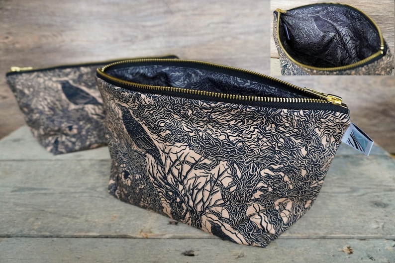 May include: Two fabric cosmetic bags with a black and tan tree and bird design. The bags have a black zipper with a gold-colored zipper pull. The interior of the bag is black. The bags are sitting on a wooden surface.