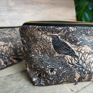 May include: Two black and tan zippered pouches with a raven and tree branch design. The raven is perched on a branch, and the background is filled with swirling lines. The zipper is gold-colored.
