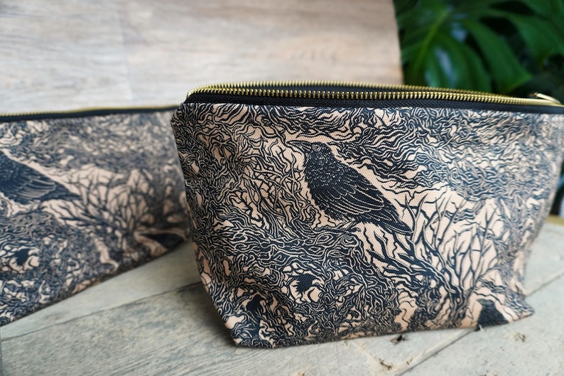 May include: Two black and tan zippered pouches with a raven and tree branch design. The raven is perched on a branch, and the background is filled with swirling lines. The zipper is gold-colored.
