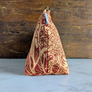May include: A triangular-shaped cosmetic bag with a repeating floral pattern in shades of red and tan. The bag has a gold zipper and a small blue tag attached. The background is a wooden surface.