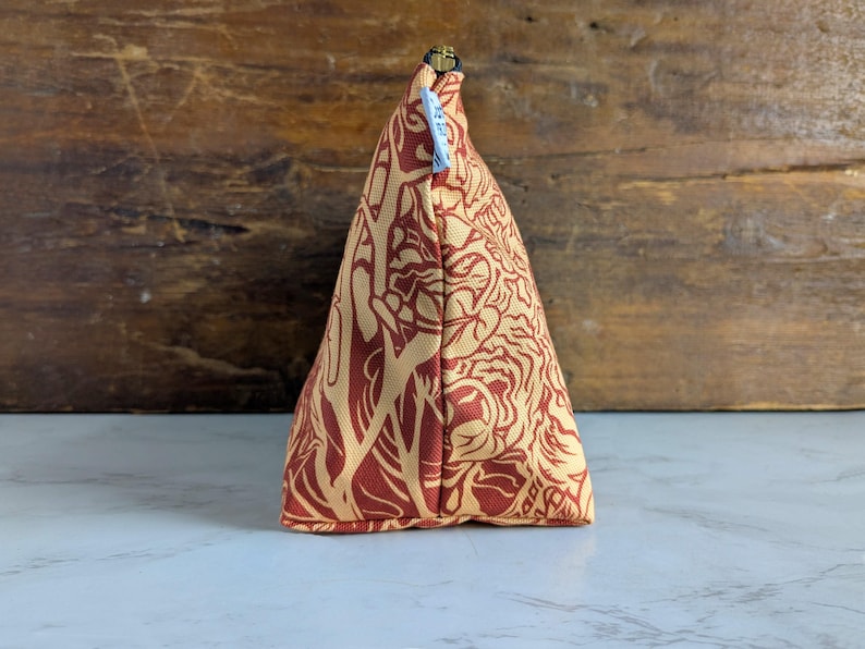May include: A triangular-shaped cosmetic bag with a repeating floral pattern in shades of red and tan. The bag has a gold zipper and a small blue tag attached. The background is a wooden surface.