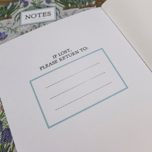 May include: A white notebook with a floral design on the cover. The cover has the word "NOTES" printed on it. The inside of the notebook has a blue border around the text "IF LOST, PLEASE RETURN TO:" and three blank lines for writing.