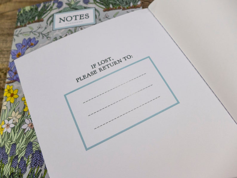 May include: A white notebook with a floral design on the cover. The cover has the word "NOTES" printed on it. The inside of the notebook has a blue border around the text "IF LOST, PLEASE RETURN TO:" and three blank lines for writing.