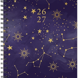 May include: Spiral-bound planner with a dark blue cover featuring gold constellations, stars, and sunburst designs. The dates "26" and "27" are visible, along with the text "JULY 2026-JUNE 2027". The planner has a metallic silver spiral binding.