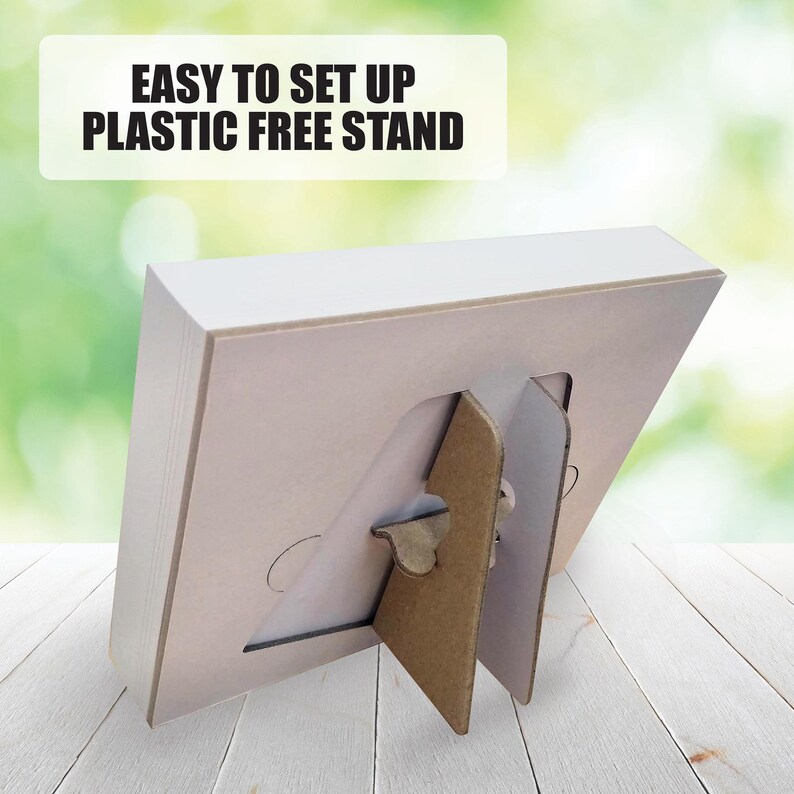May include: A white, angled cardboard stand with a brown support. The stand is on a white wooden surface. Text at the top reads "EASY TO SET UP PLASTIC FREE STAND". The background is a blurred green and white.
