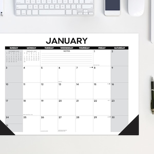 May include: A white desk calendar pad for the month of January with black text and grid lines. The calendar features a notes section, small reference calendars for December and February, and black corner protectors on the bottom left and right.