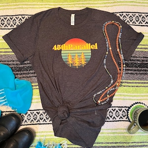 Könnte beinhalten: Ein dunkelgraues T-Shirt mit einer Retro-Sonnenuntergangsgrafik und dem Text "45th Parallel". Das Shirt liegt auf einer gestreiften Decke mit einer Perlenkette und einer Tasse.