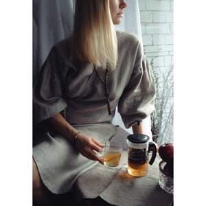 May include: A woman wearing a gray linen dress sits by a window, holding a glass of tea. A glass teapot with a black lid and handle sits on a table in front of her. The teapot is filled with tea and has a strainer inside. There are red apples in a bowl on the table.