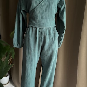 May include: A teal two-piece outfit on a mannequin. The top is a wrap-style blouse with long sleeves. The trousers are loose-fitting with elasticated ankles. The fabric has a textured, crinkled appearance.