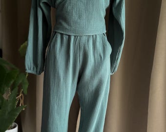 Puede incluir: Un conjunto de dos piezas de color verde azulado exhibido en un maniquí. La parte superior es una blusa cruzada con mangas largas. Los pantalones son holgados con tobillos elásticos. La tela tiene una apariencia texturizada y arrugada.