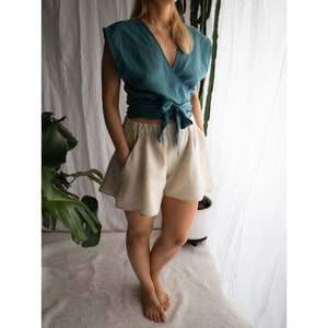 May include: A teal wrap top with short sleeves and a tie-front closure paired with beige high-waisted shorts featuring an elastic waistband and side pockets. The ensemble consists of a solid-colored teal blouse and neutral-toned loose-fitting shorts.