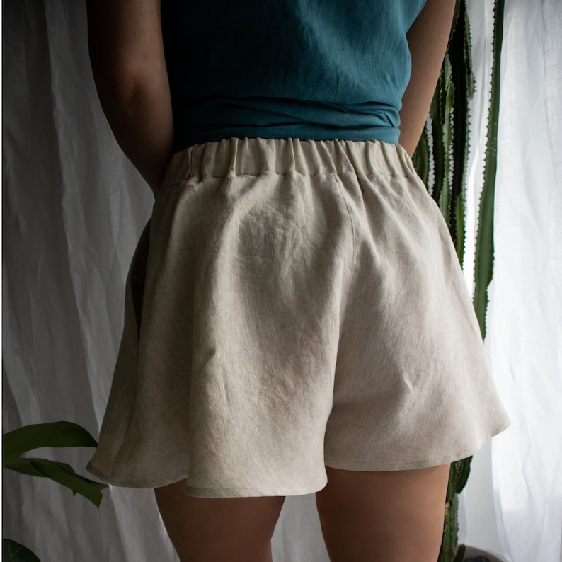 May include: Beige shorts featuring a high-waisted design with an elasticized waistband and a flared, loose-fitting silhouette. The shorts are crafted from a textured fabric and include side pockets, providing a relaxed fit that sits at the upper thighs.