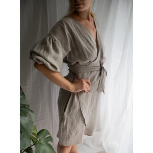 May include: A light brown linen wrap dress with a V-neckline and puffed sleeves. The dress has a tie at the waist and a flowing skirt.