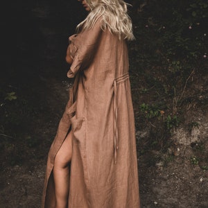 May include: A woman wearing a long, brown linen robe with a tie at the waist. The robe has a slit up the side, revealing her leg. She is standing in a forest setting.